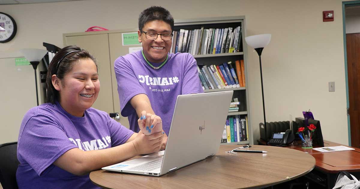 两个穿着SJC t恤的学生看着电脑，面带微笑